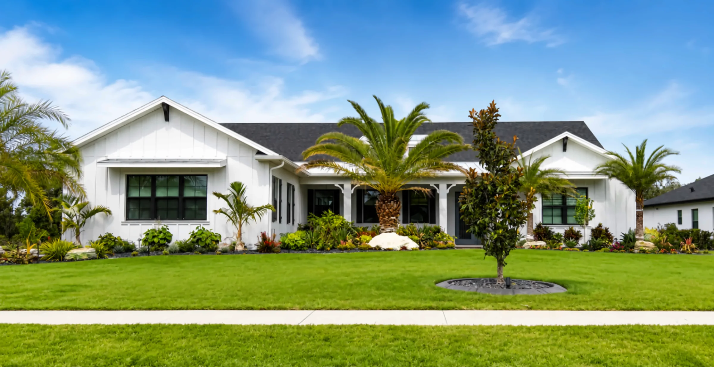 White farmhouse-style home with a finished front landscape of palms, tropical beds, boulders and a fresh lawn
